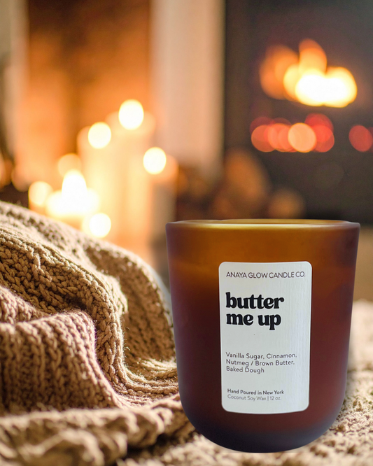 Hand-poured Butter Me Up candle by Anaya Glow Candle Co., set in a warm, cozy space with soft lighting and candles glowing in the background.